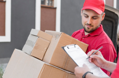 Two delivery workers in red uniforms handling a stack of cardboard boxes beside a van, one filling out a form on a clipboard.