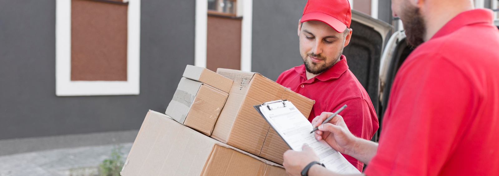 Two delivery workers in red uniforms handling a stack of cardboard boxes beside a van, one filling out a form on a clipboard.