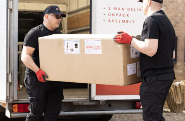 Two workers in black uniforms carry a large cardboard box from a truck, wearing red gloves, beside a 'Higher Jump Logistics' sign on the trailer.